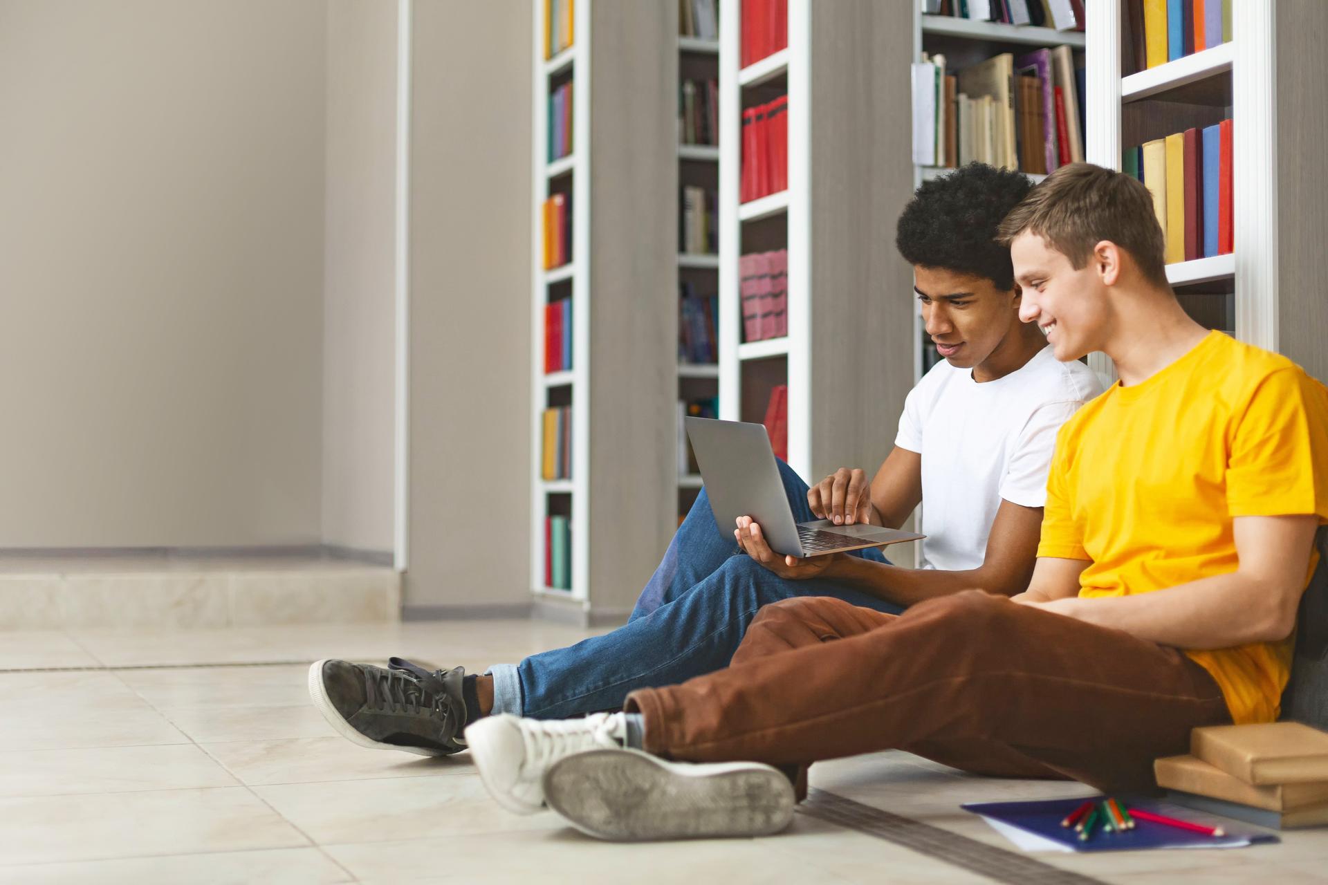 zwei junge Männer sitzen vor Bücherregalen auf dem Boden und lernen