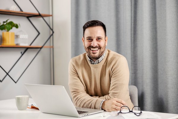 Ein freundlich lächelnder Mann arbeitet an seinem Laptop und hält seine Brille in einer Hand