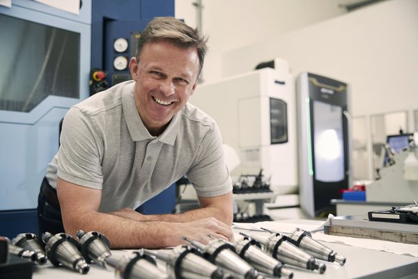 ein Meister steht lächelnd vor einem Tisch mich technischen Bauteilen